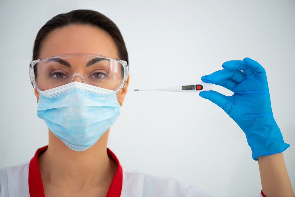 A healthcare worker wearing a mask, goggles, and blue gloves holds a digital thermometer, showing a reading.